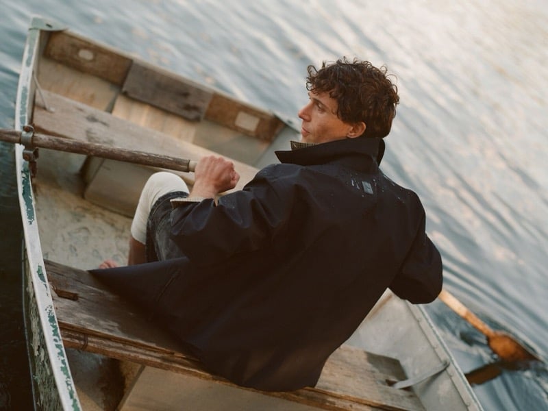 William Beck rows a wooden boat across the lake in a Dior shell jacket dotted with water droplets