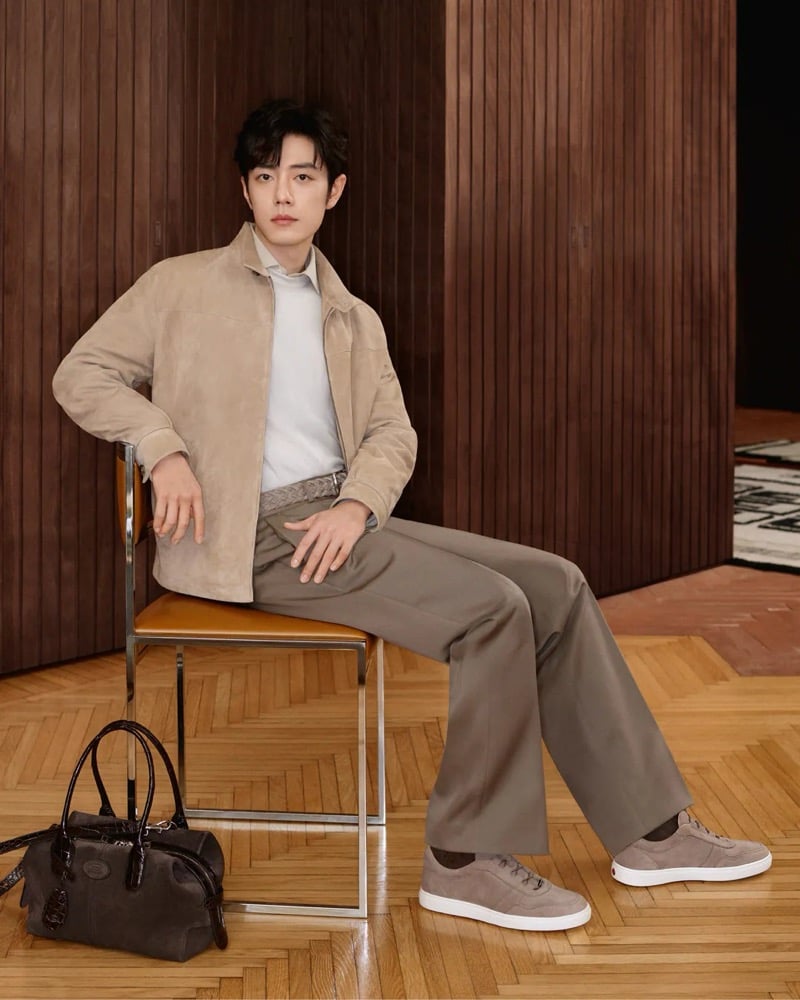 Xiao Zhan sits on a leather-topped chair in a taupe suede blouson and white shirt with wide pleated trousers and suede low-top sneakers, a dark brown suede Di Bag holdall on the parquet floor beside him