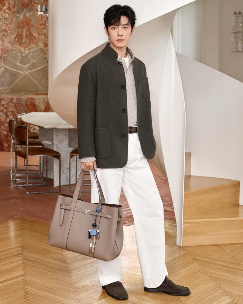 Xiao Zhan stands beside a spiral staircase in a marble-walled villa wearing a stand-collar wool blazer over a marled knit with a leather belt and light wide-leg trousers, holding a greige leather Di Bag tote with a blue bag charm