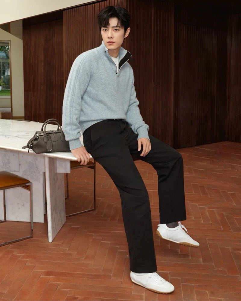Xiao Zhan leans against a marble table in a half-zip raglan sweater over a white tee with dark trousers and white sneakers, a small brown suede Di Bag on the table