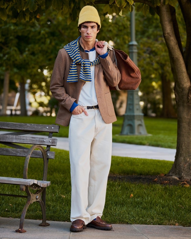 Duncan Yair wears a brown blazer, white henley, pale trousers, and gold beanie, with a striped top draped over his shoulders.