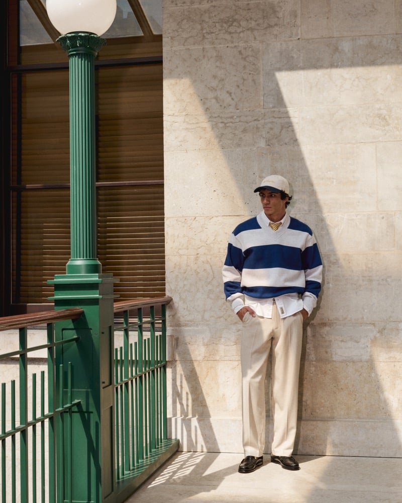Duncan Yair wears a navy-and-cream striped V-neck sweatshirt with a white shirt, tie, pale trousers, and baseball cap.