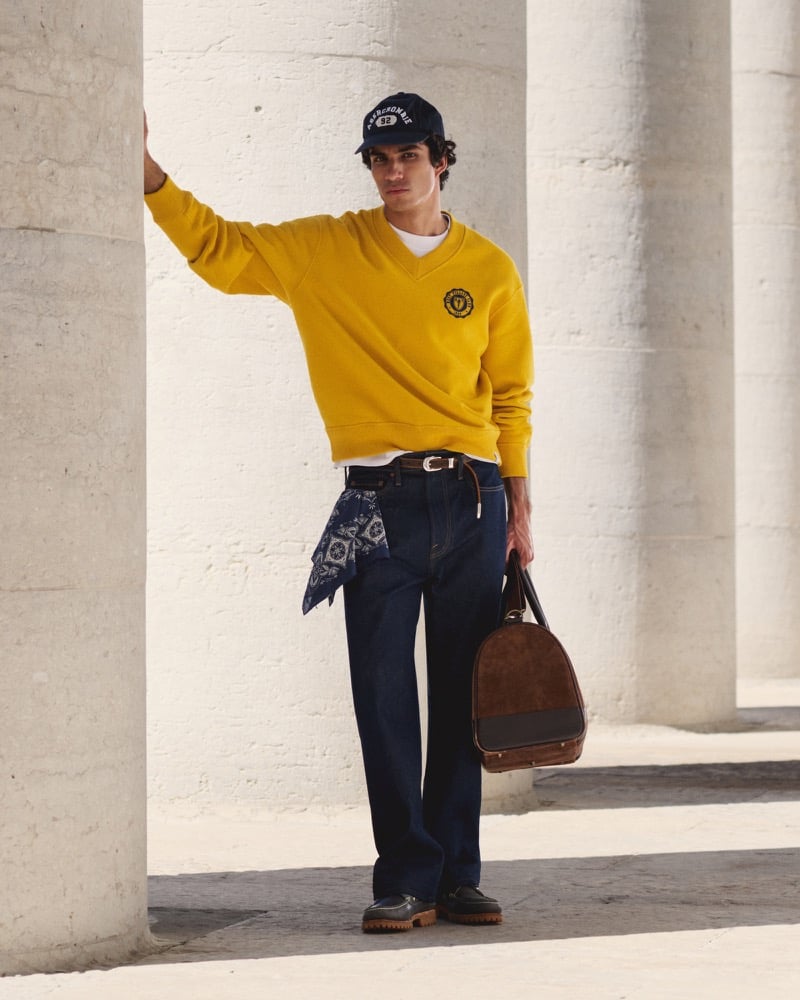 Duncan Yair wears a yellow collegiate V-neck sweatshirt, dark trousers, and navy baseball cap, carrying a brown bag.