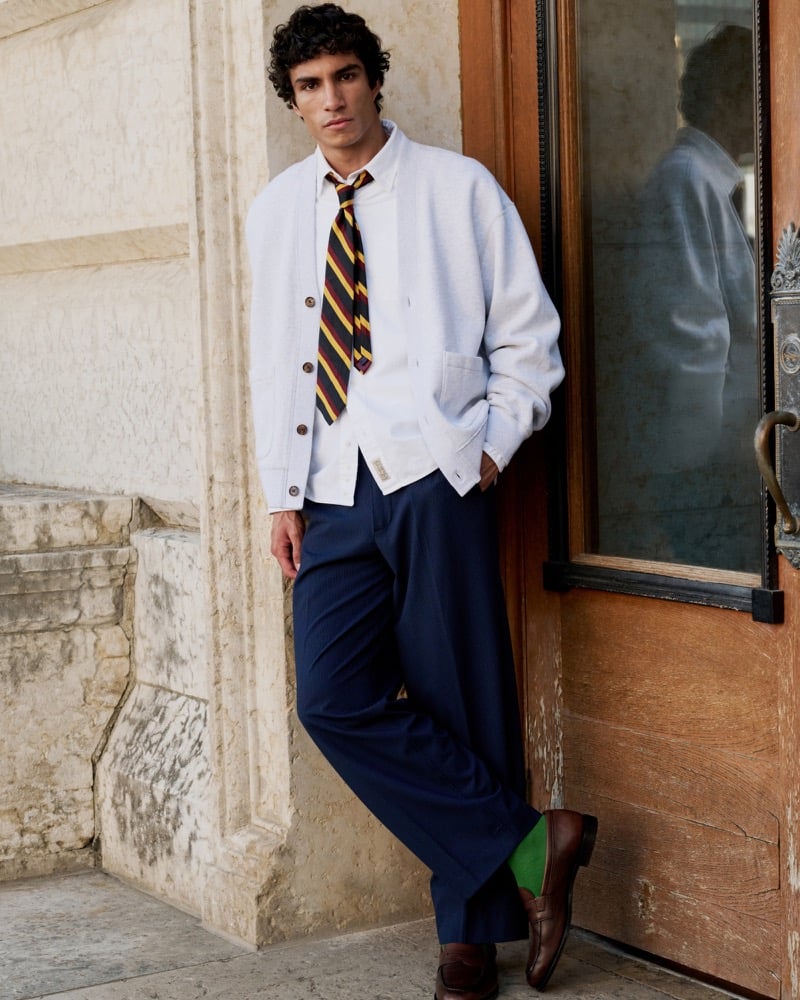 Duncan Yair wears a pale cardigan, striped tie, and navy trousers beside a wooden doorway.