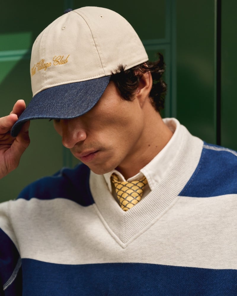 Duncan Yair tops a striped sweatshirt, white shirt, and patterned tie with Abercrombie & Fitch’s West Village Club baseball hat.