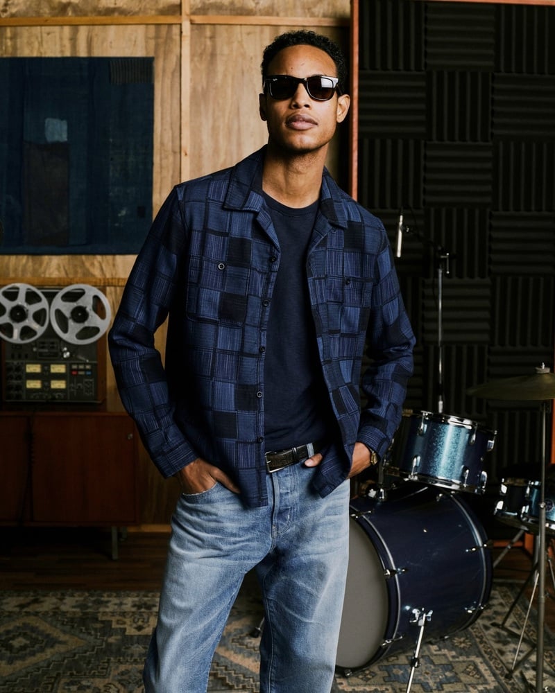 Conrad Bromfield wears sunglasses and a blue patchwork jacket with light-wash jeans.