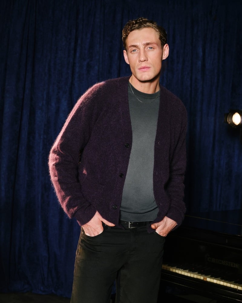 Aubrey O'Mahony wears a deep purple cardigan over a gray top against blue curtains.