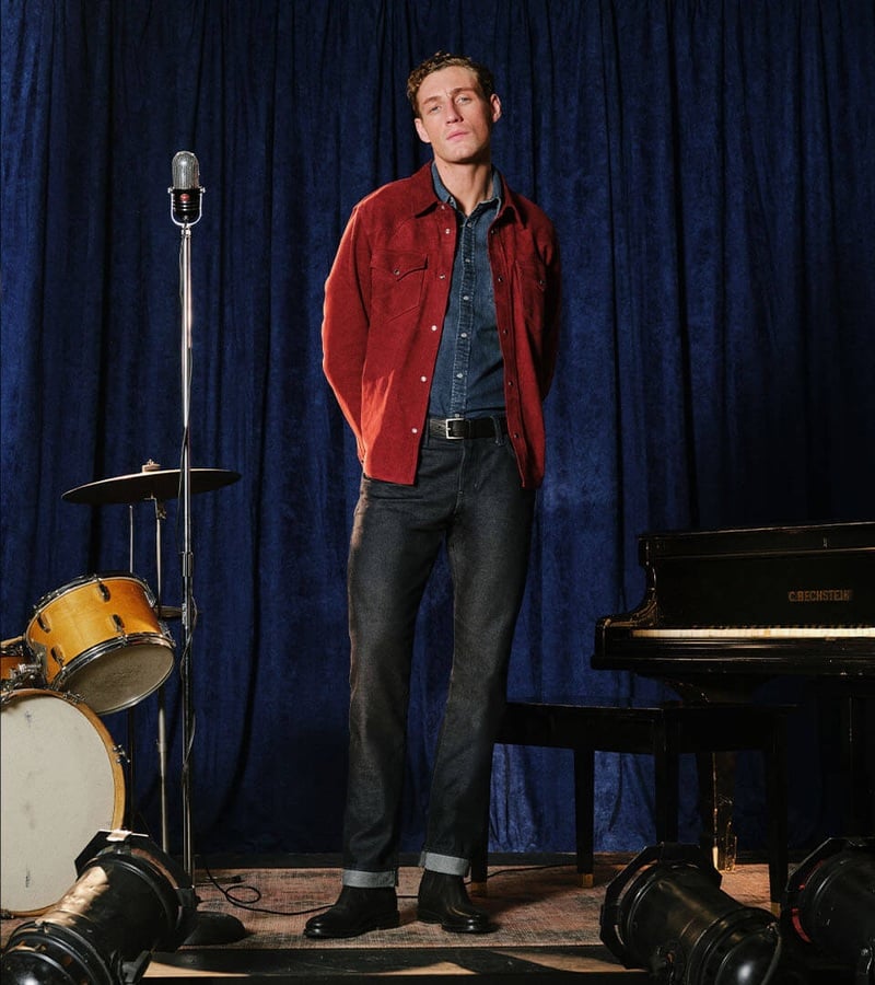 Aubrey O'Mahony wears a red overshirt with a denim shirt and cuffed jeans beside a microphone.