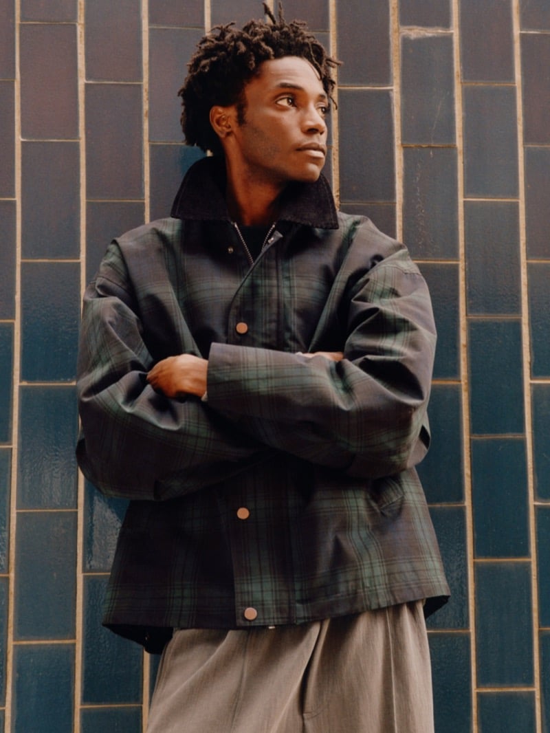 Ahmed Richards standing with arms crossed against a wall of glazed tiles in an oversized green plaid snap-front jacket with a dark corduroy collar and wide trousers