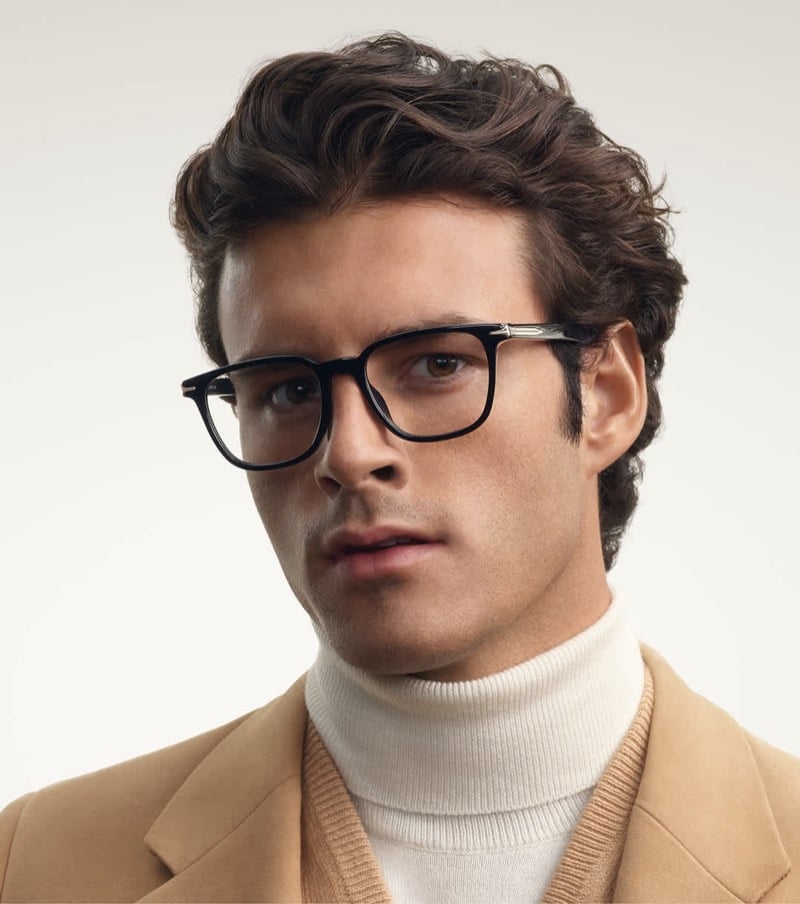 Aiden Stonehouse wears black rectangular optical frames with metal temple accents, camel tailoring, and layered knitwear.