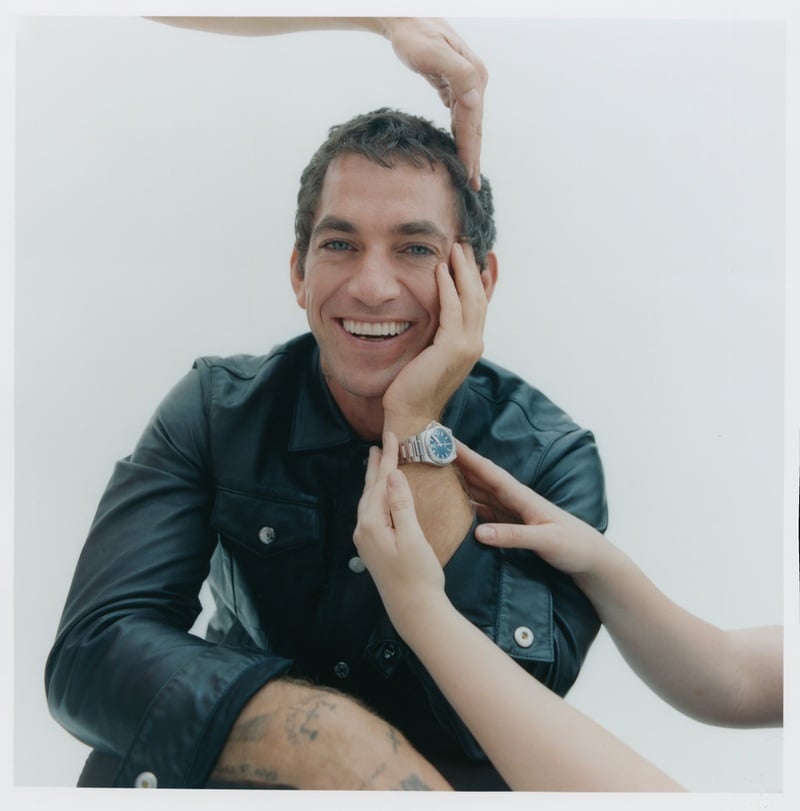 Alex Fine smiles in a leather jacket while hands adjust his hair and wristwatch.