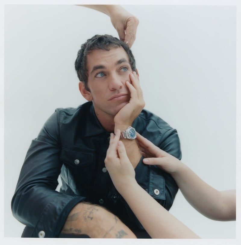 Alex Fine looks upward in a leather jacket as hands adjust his hair and IWC watch.
