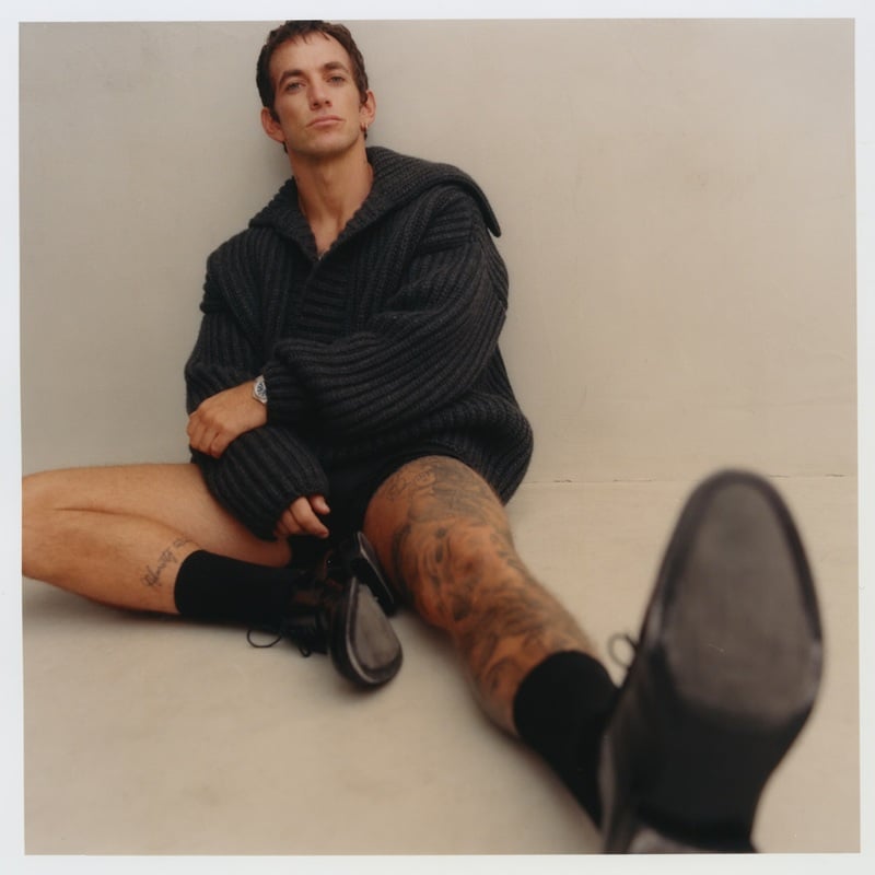 Alex Fine sits with his legs extended in a dark Dolce & Gabbana sweater and shoes.