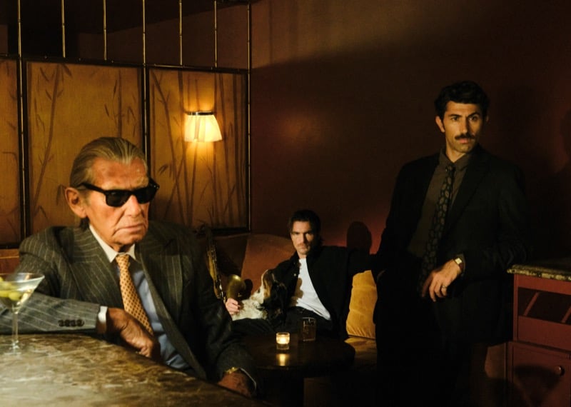 Three men in a dimly lit lounge: one in sunglasses and a pinstripe suit at a marble table, one on a sofa with a dog, and one standing in a dark suit
