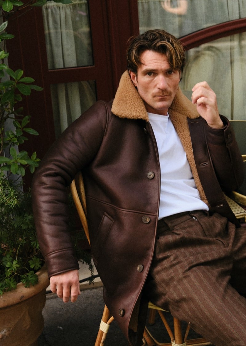 Anthony L. seated on a rattan chair outside a café in a brown shearling-lined leather jacket and pinstripe trousers