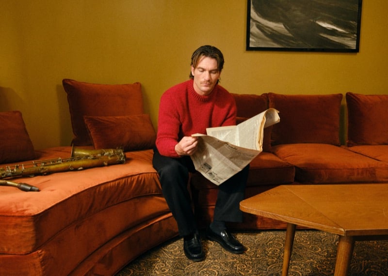 Anthony L. reading a newspaper on a velvet sofa in a red sweater and loafers, a saxophone lying on the cushions beside him