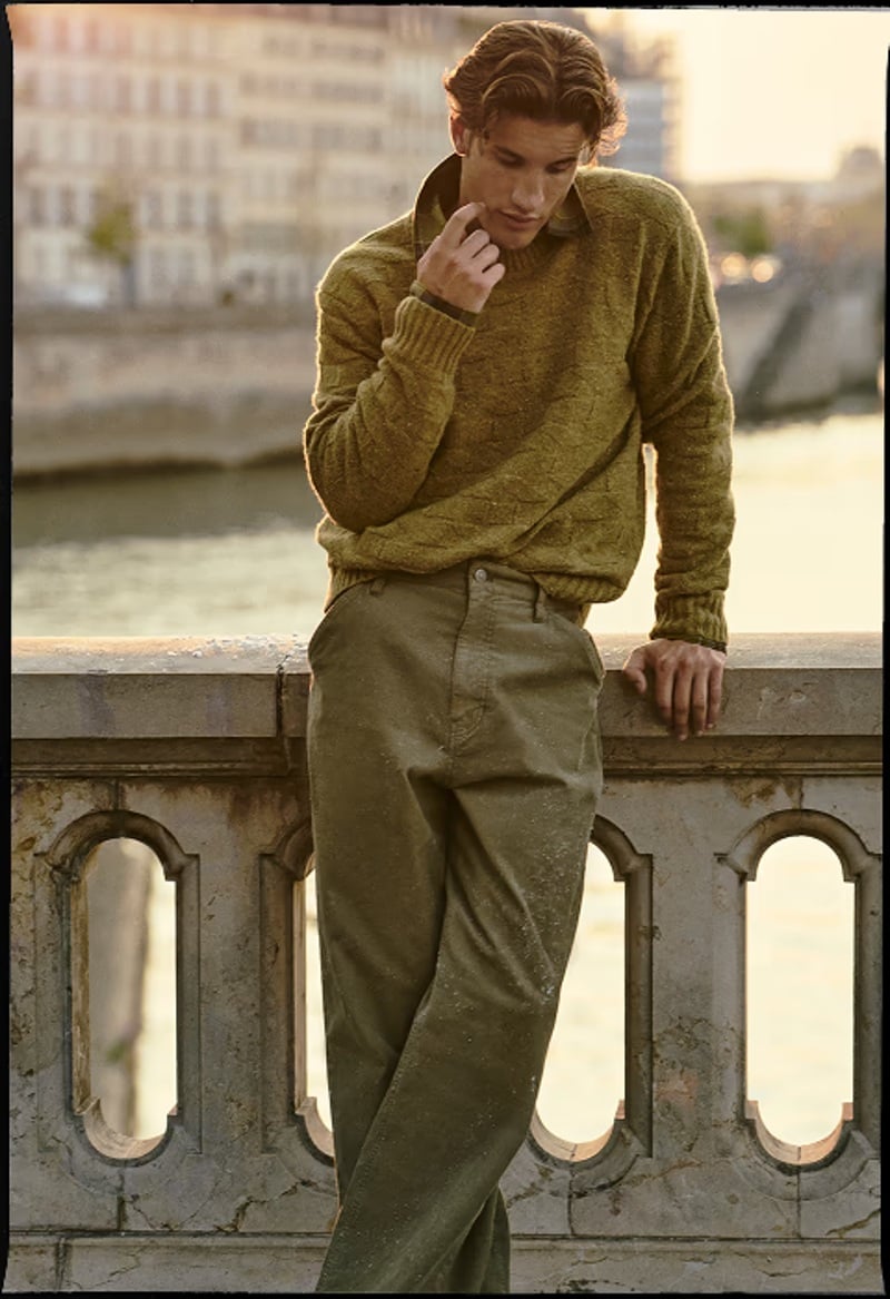 Max Knott wears an olive cable-knit sweater and loose trousers beside a Paris balustrade.