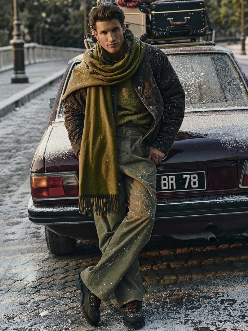 Max Knott wears a jacket and olive scarf beside a luggage-topped car.