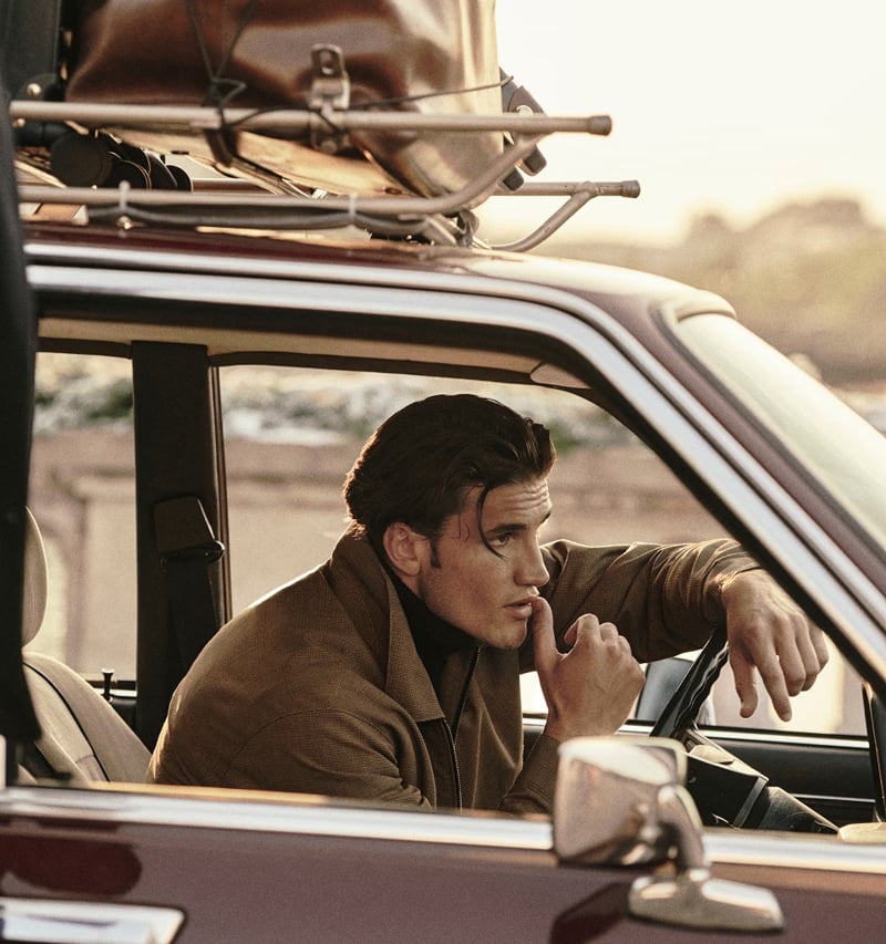 Max Knott sits at the wheel of a luggage-topped car in a brown jacket.