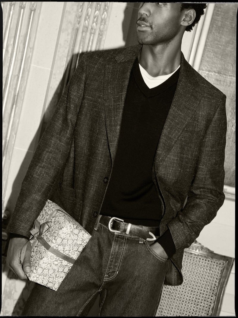 Halid Nuhu wears a checked blazer, dark V-neck sweater, and jeans while holding a wrapped gift.