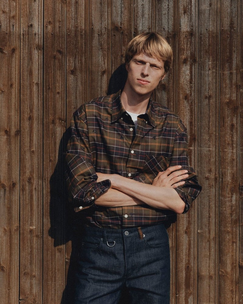 Joseph Uyttenhove wearing a tartan shirt tucked into dark jeans against a wooden wall for Barbour x A.P.C.
