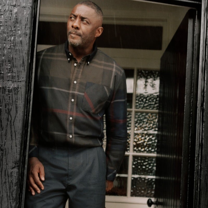 Idris Elba wears a tartan shirt tucked into pleated trousers at a pub doorway for Barbour.