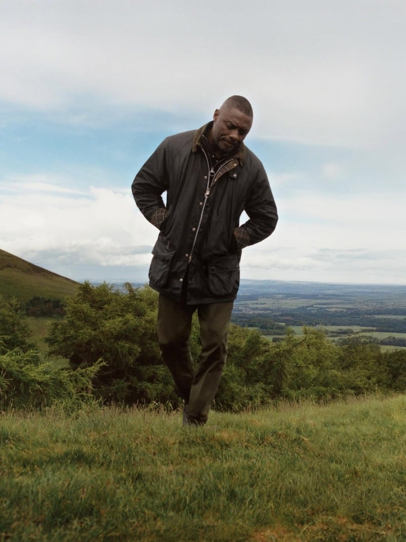 Idris Elba walks across a grassy hillside in a buttoned Barbour jacket.