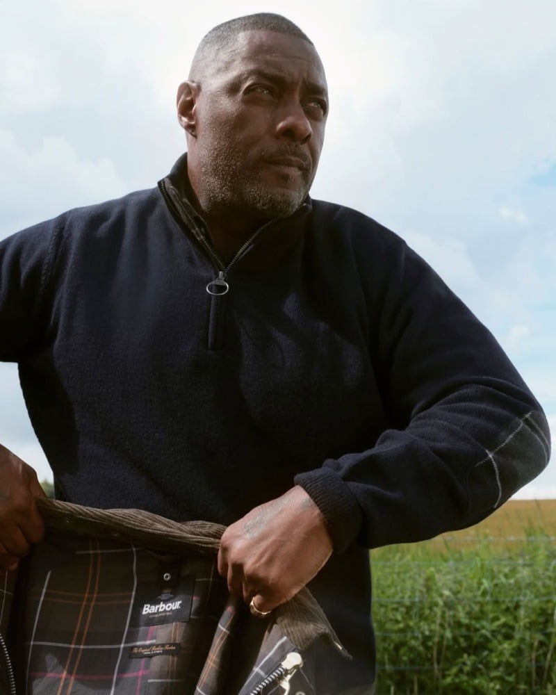 Idris Elba wears a quarter-zip sweater while holding a tartan-lined Barbour jacket.