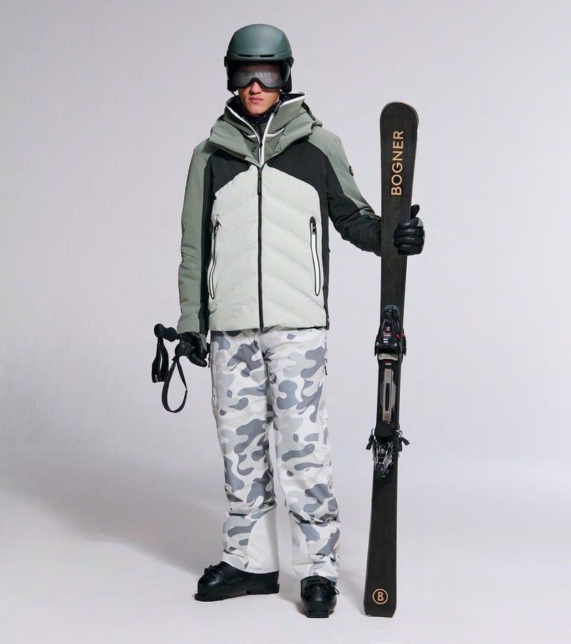 A model holds skis wearing a gray and black jacket with camouflage ski trousers. Bogner fall-winter 2026 collection.