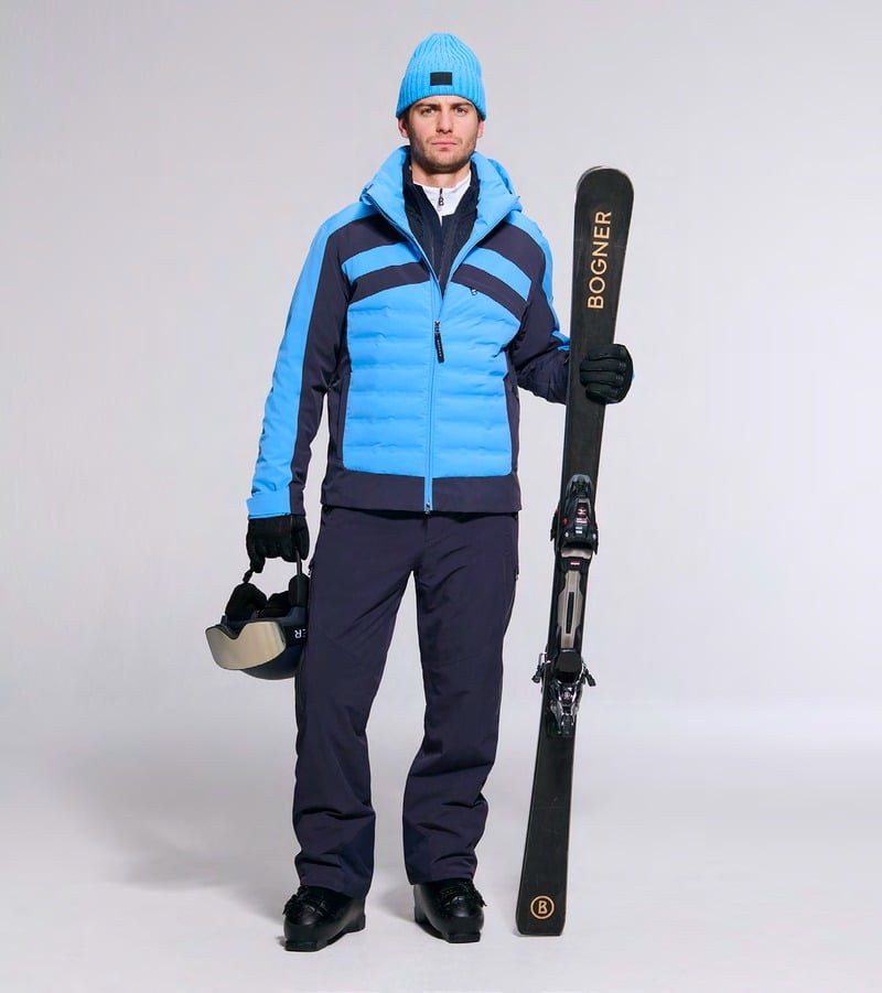 A model holds skis wearing a blue and black jacket with navy trousers and a blue beanie. Bogner fall-winter 2026 collection.