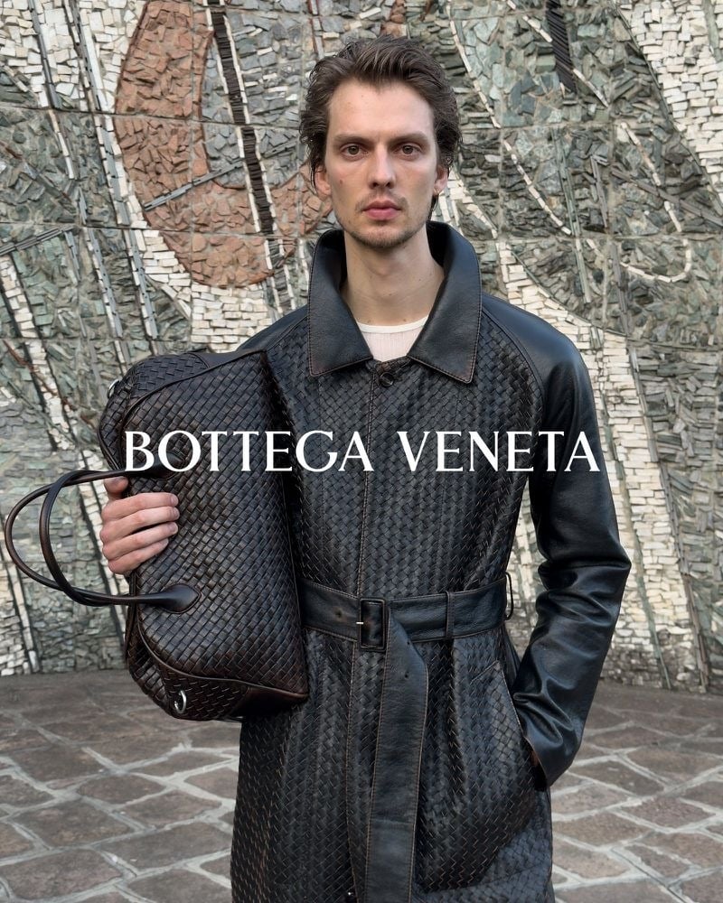 Leon Dame wearing a black belted coat with woven panels and holding a dark woven bag.