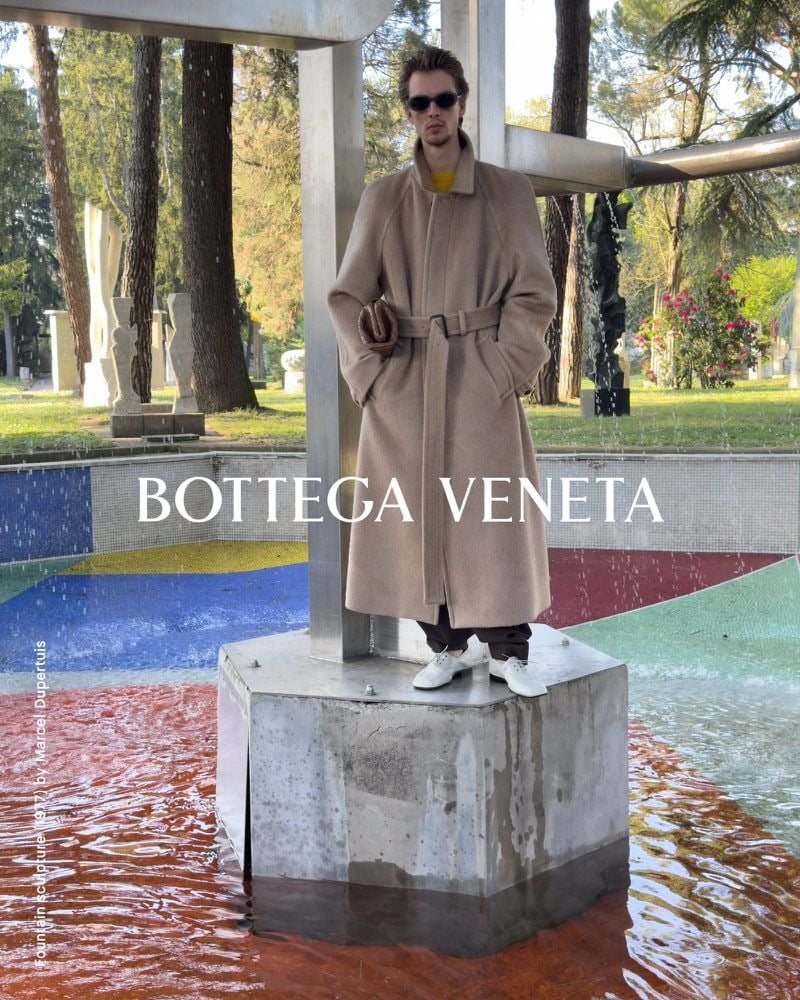 Leon Dame in sunglasses and a belted camel coat standing on a concrete pedestal above a reflecting pool.