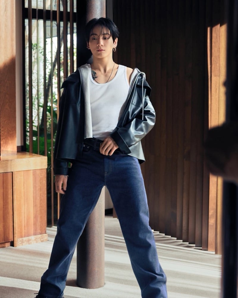 Jung Kook poses beside a column in a white tank, black leather jacket, and blue jeans behind the scenes for Calvin Klein.