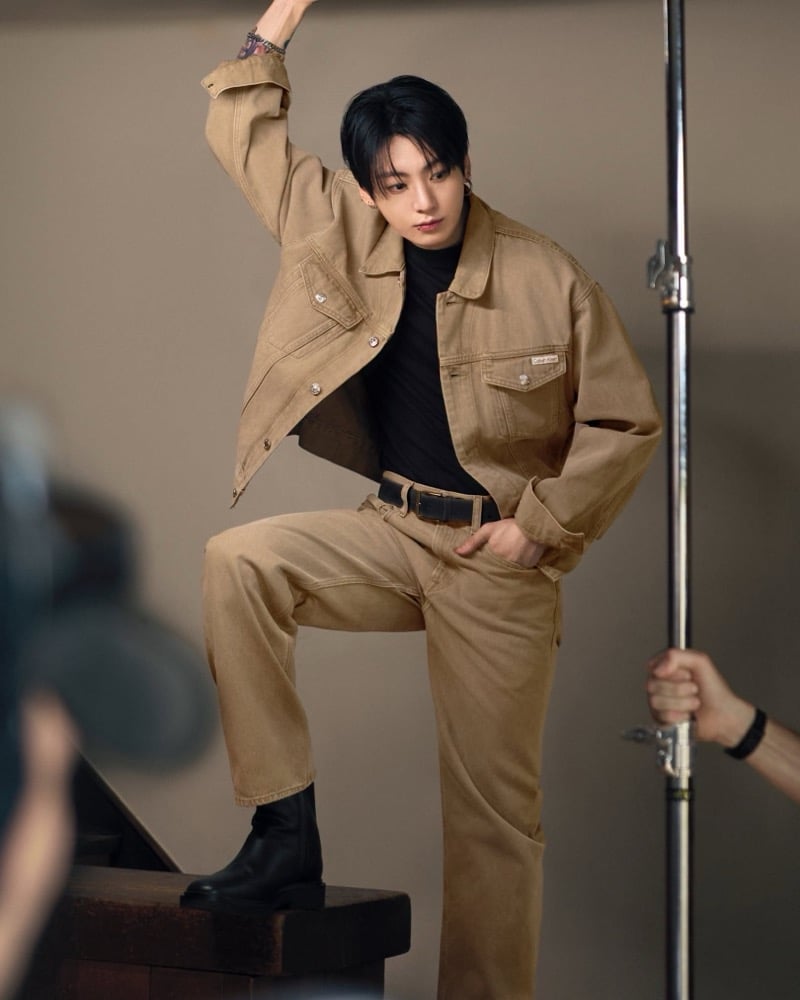 Jung Kook raises one arm and rests a boot on a bench in a tan jacket and matching pants on the Calvin Klein set.