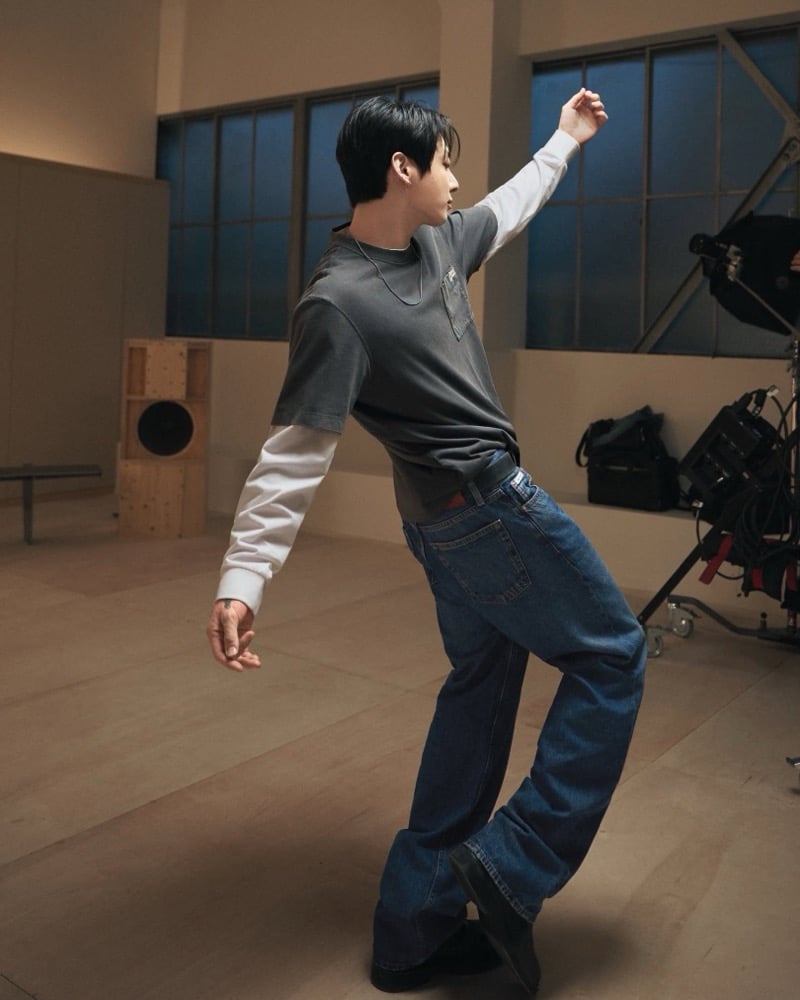 Jung Kook leans back in layered gray and white tees and blue jeans beside studio equipment on the Calvin Klein set.