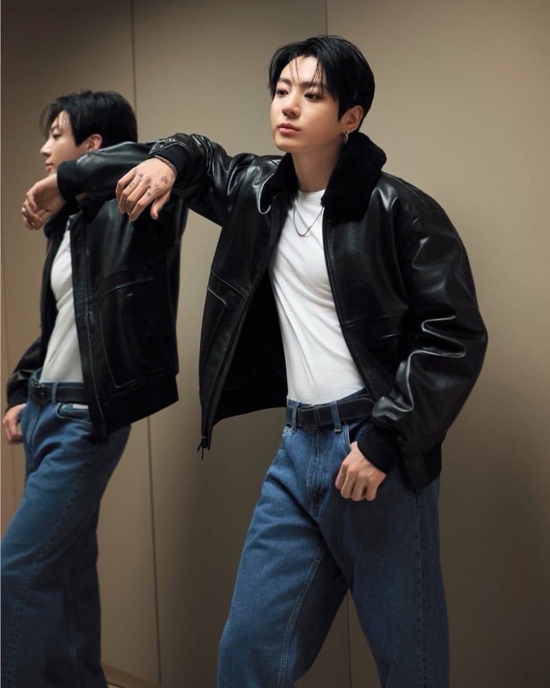 Jung Kook rests his arm against a mirror in a black leather jacket, white tank, and blue jeans behind the scenes for Calvin Klein.