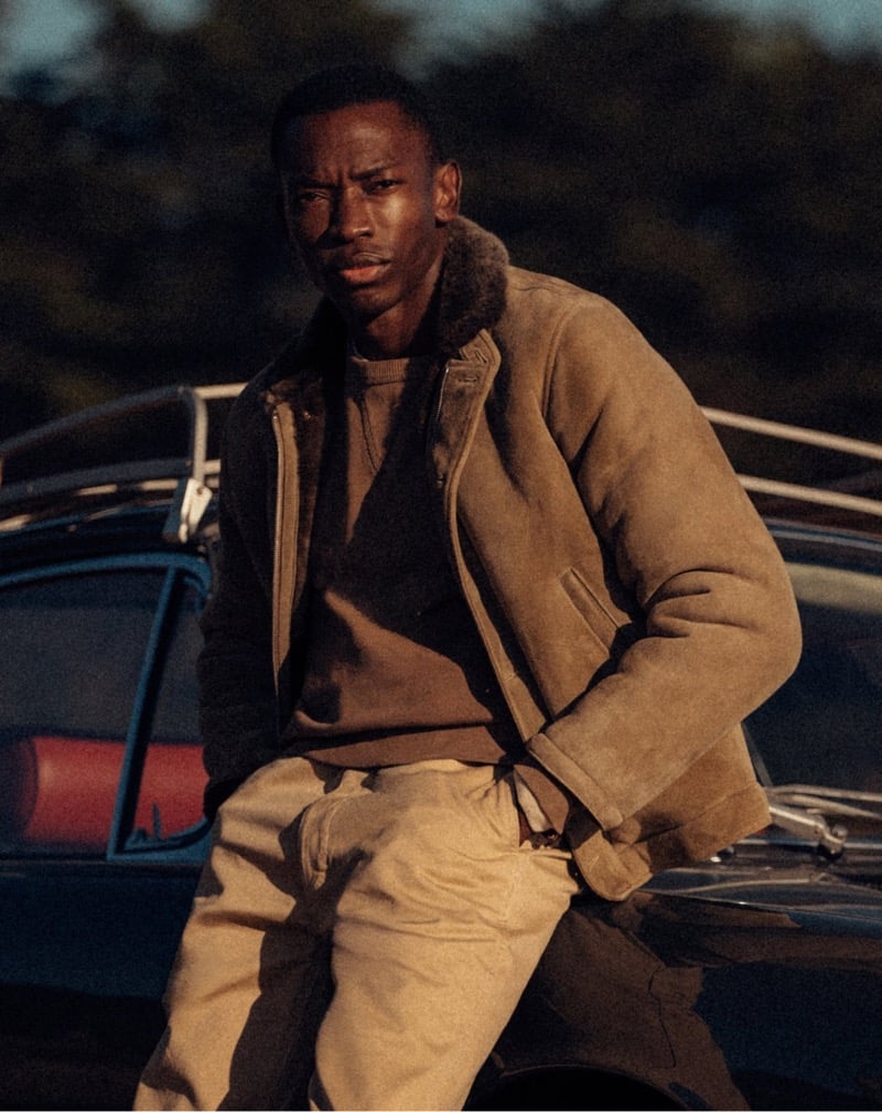 Akin Jaiye leans against a vintage car in a shearling-lined deck jacket and chinos.