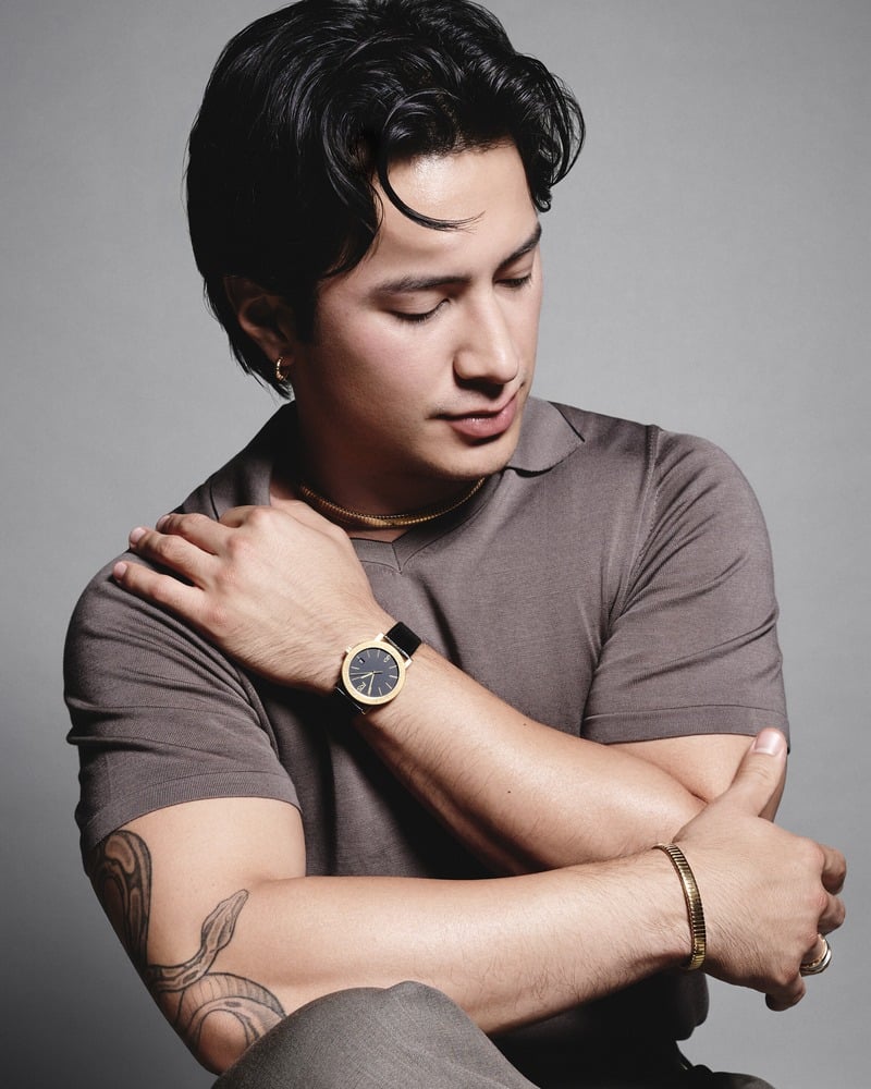 Hudson Williams looks down at his Bvlgari watch, wearing a polo, necklace, bracelets, rings, and earrings.