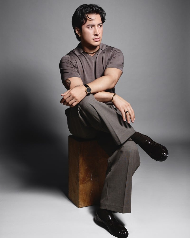 Hudson Williams sits on a wooden block in a polo and tailored trousers with Bvlgari jewelry and a watch.