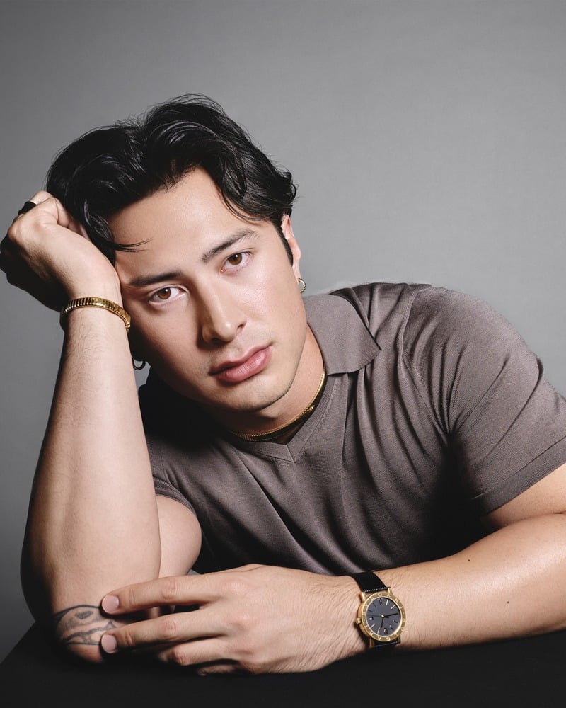 Hudson Williams rests his head on his hand at a table, wearing Bvlgari jewelry and a watch.