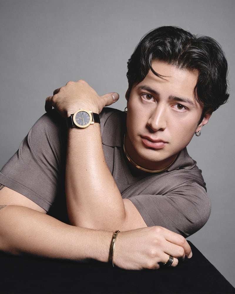 Hudson Williams holds his wrist beside his face, showing a Bvlgari dark-dial watch, necklace, earrings, bracelet, and rings.
