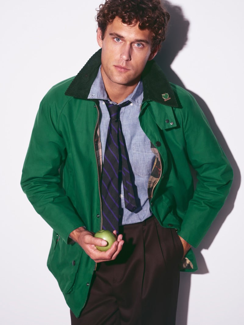 Conor Fay in a green waxed Beaufort jacket over a chambray shirt and striped tie, holding a green apple in one hand
