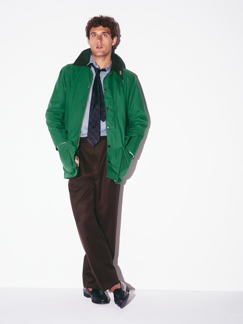 Conor Fay standing full-length in a green Beaufort jacket with a dark corduroy collar, a chambray shirt, a striped tie, wide pleated brown trousers, and loafers