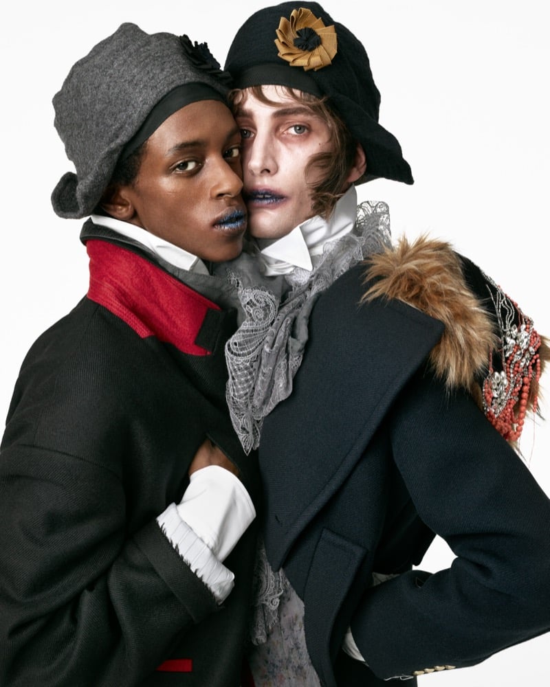 Craig Shimirmana in a gray felt hat and wool coat with a red collar embracing Thomas Riguelle in a black hat, lace jabot, and fur-trimmed beaded coat