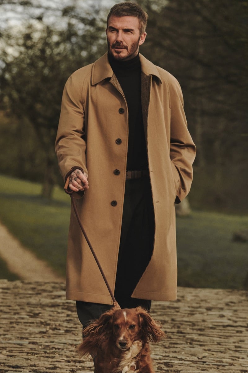 David Beckham walking a spaniel on a leash along a stone path, wearing a long camel overcoat with a dark turtleneck and trousers