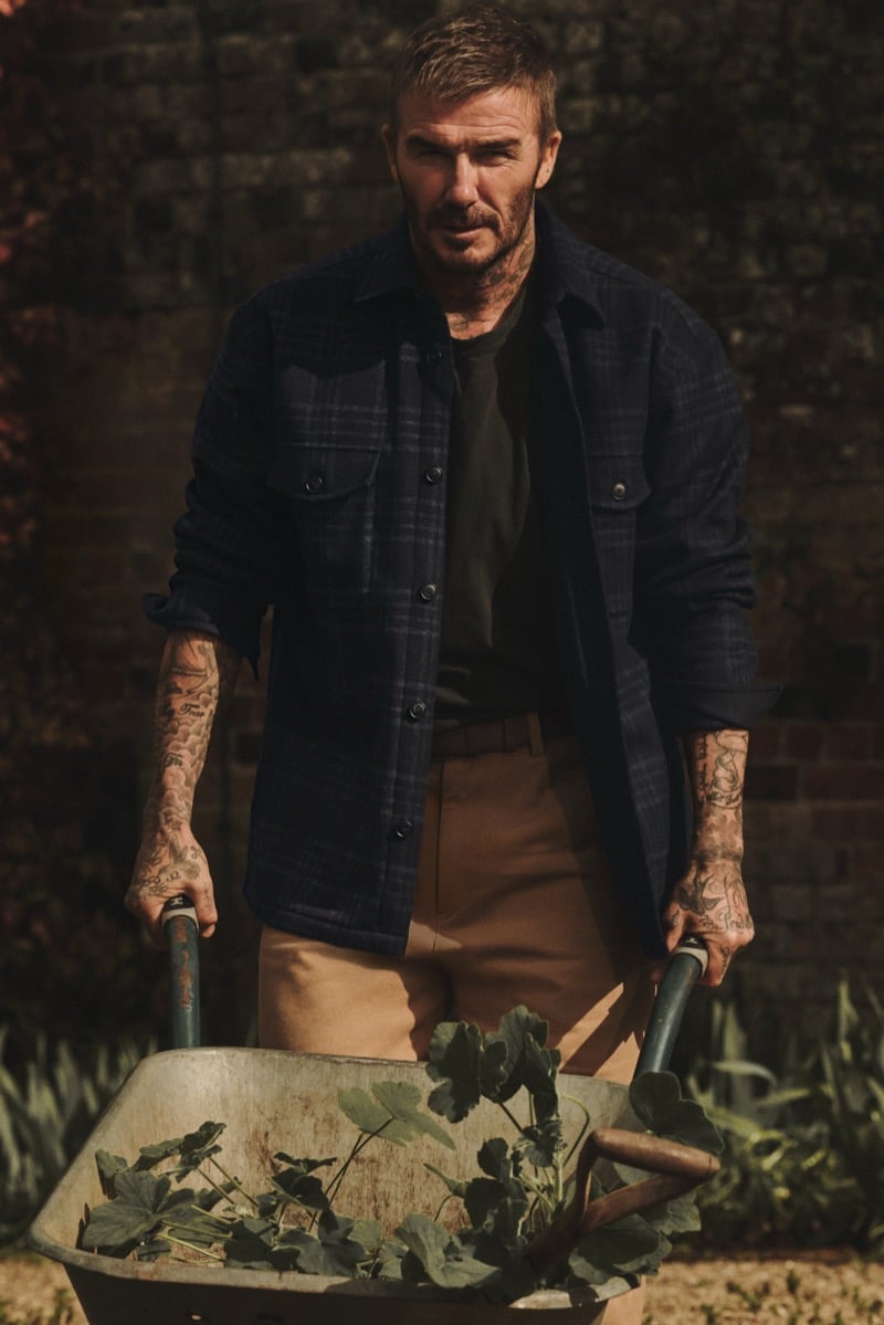 David Beckham pushing a wheelbarrow of garden leaves in front of a brick wall, wearing a dark checked overshirt with rolled sleeves, a dark tee, and tan trousers