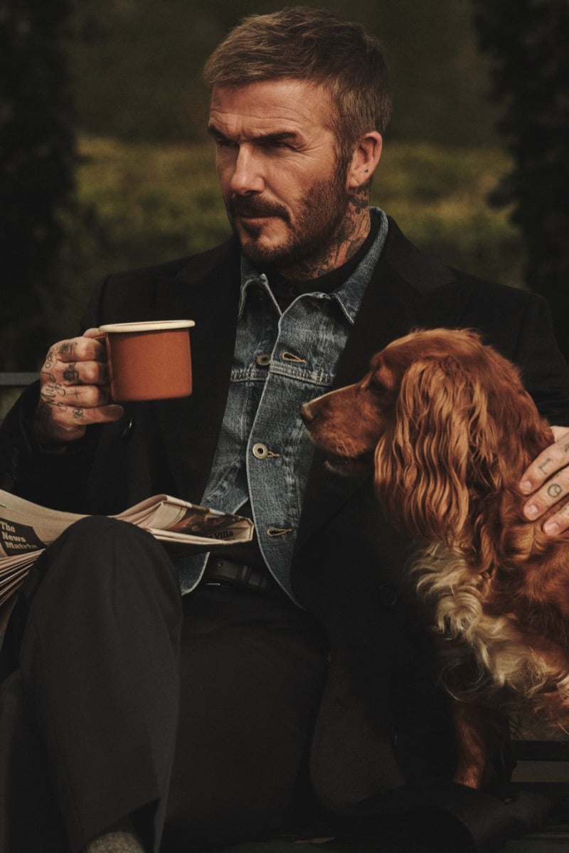 David Beckham seated outdoors with a newspaper on his lap and an enamel mug in hand, a denim jacket under a dark tailored coat, one hand resting on a spaniel