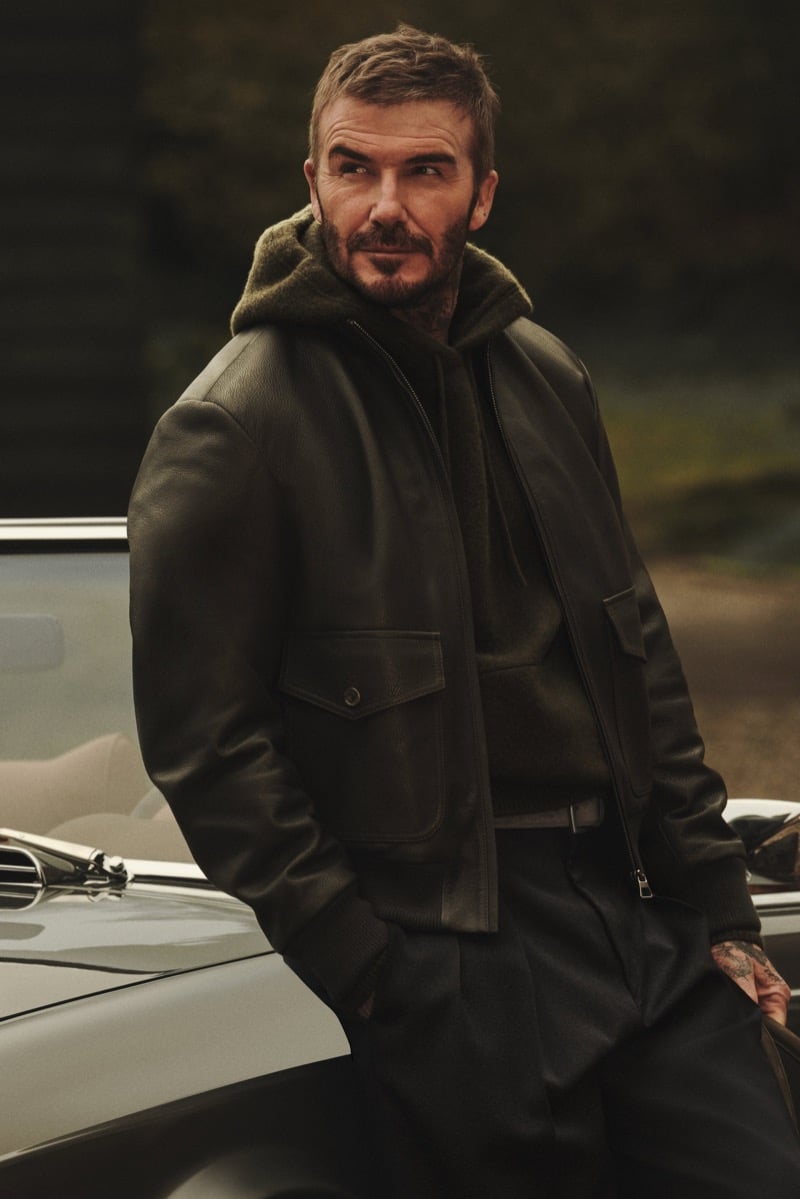 David Beckham leaning against a vintage convertible with a hand in his pocket, wearing a dark leather bomber jacket with flap pockets over a hooded knit