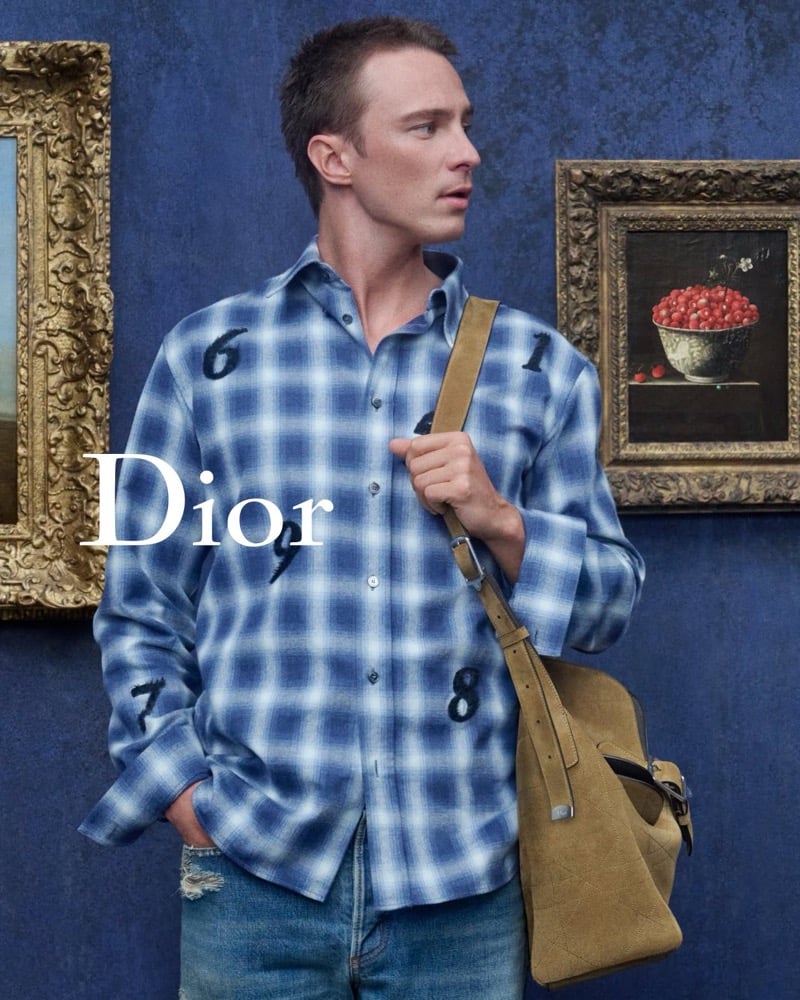Drew Starkey in a blue plaid shirt and faded jeans with a tan shoulder bag beside framed paintings at LACMA.