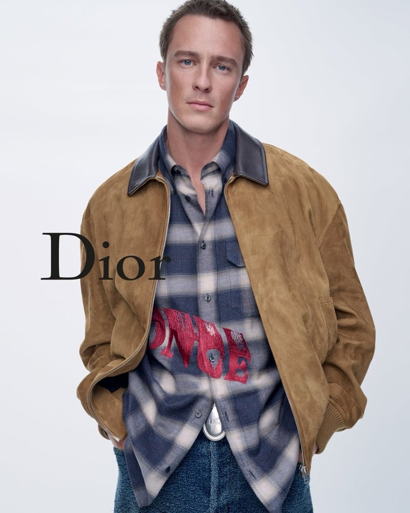 Drew Starkey in a tan jacket with a dark collar over a gray and blue plaid shirt.